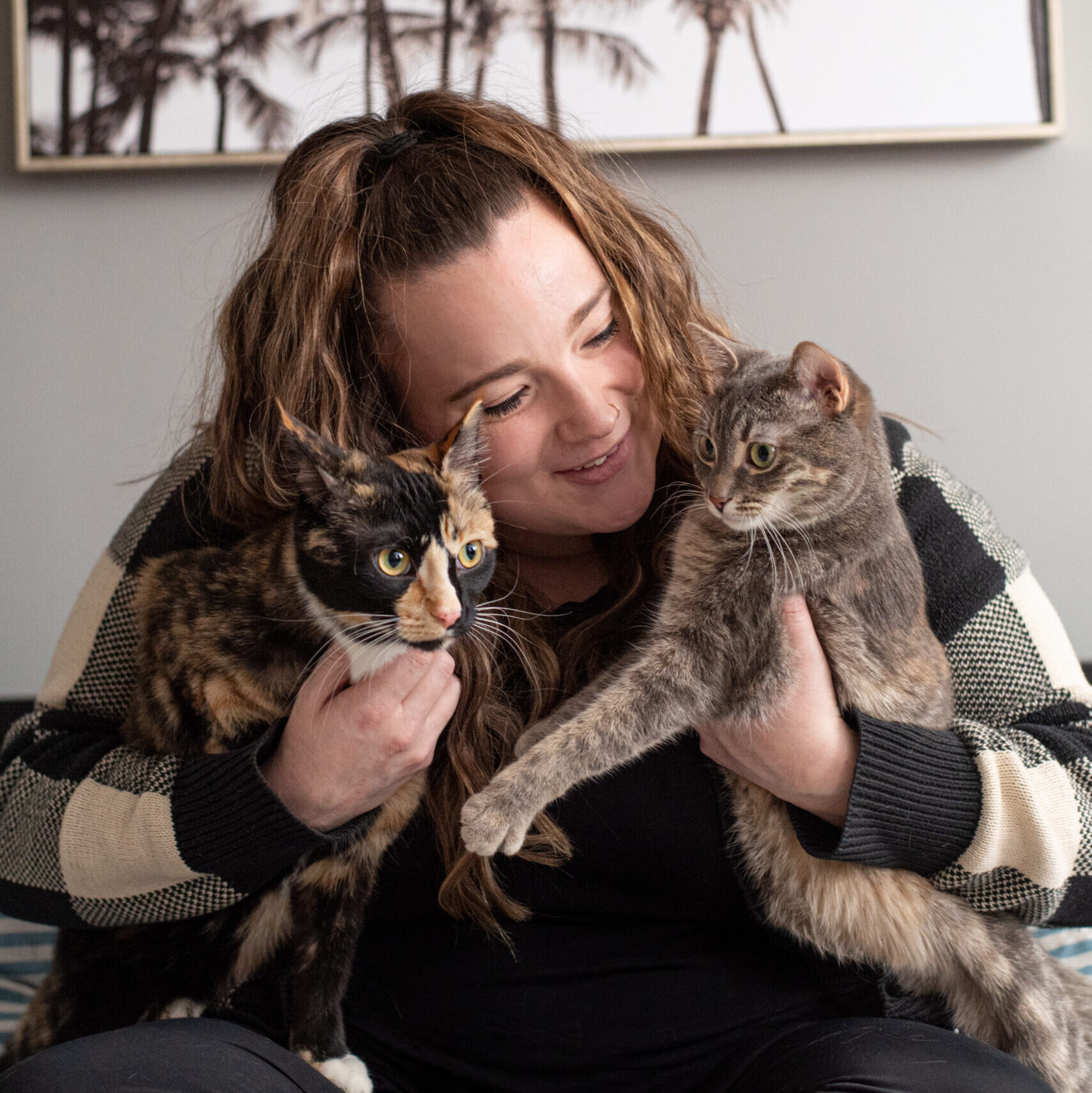 A woman with long wavy hair sitting cross-legged on a bed lovingly holding two multi-colored cats