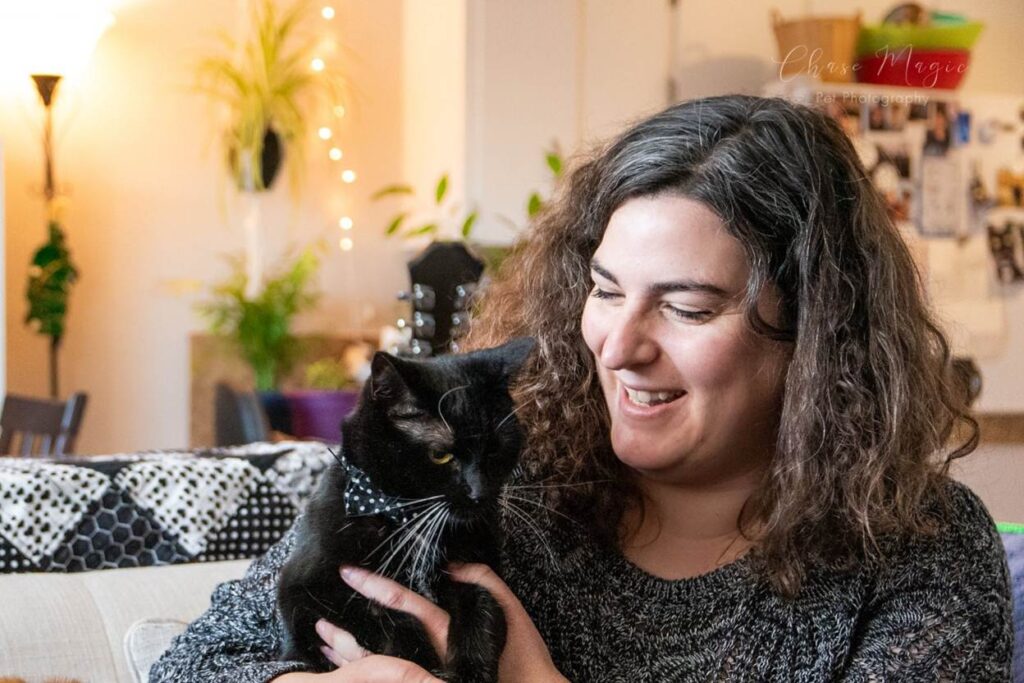 A woman with brown curly hair sitting on a couch looks down lovingly at her black cat. In the background are plants and a kitchen table with white christmas lights 