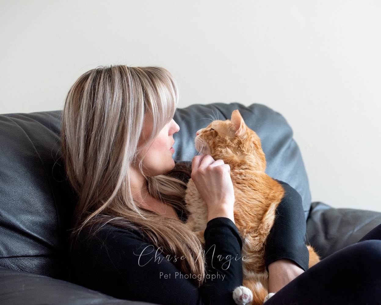 A woman with long blonde hair smiles at an orange tabby cat sitting on her chest. She is sitting on a black leather couch, wearing a black top, and the cat looks at her affectionately. The photos show warm, natural lighting, emphasizing the close bond between the woman and her pet in Calgary